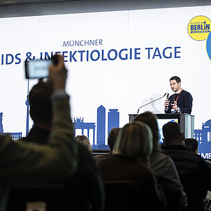 Referent auf der Bühne während seines Vortrags bei den 20. Münchner AIDS- und Infektiologie-Tagen 2026 in Berlin. Teilnehmerschaft im Vordergrund.